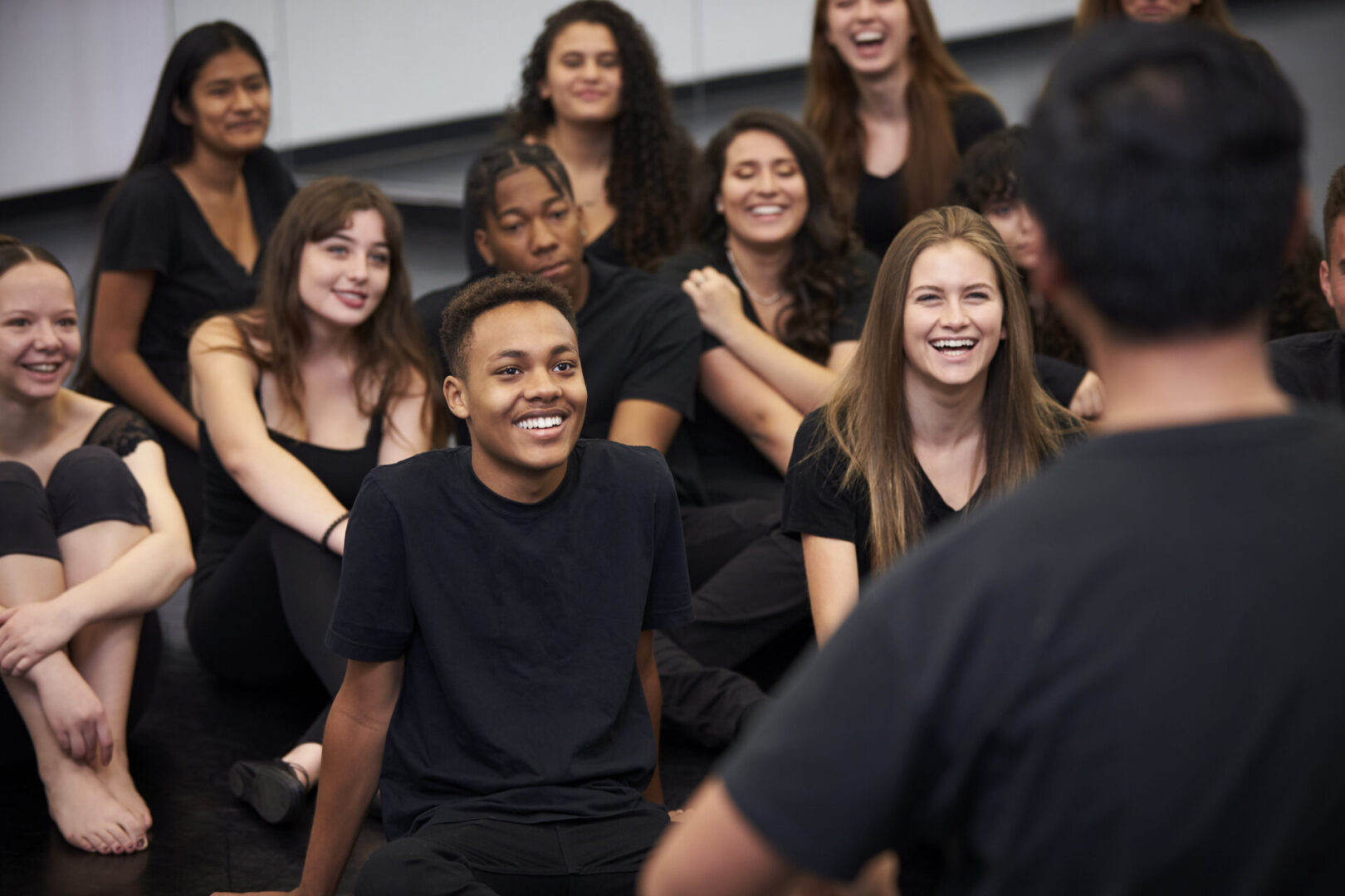 Teacher At Performing Arts School Talking To Students Sitting On Floor In Rehearsal Studio Group of people sitting and smiling together.