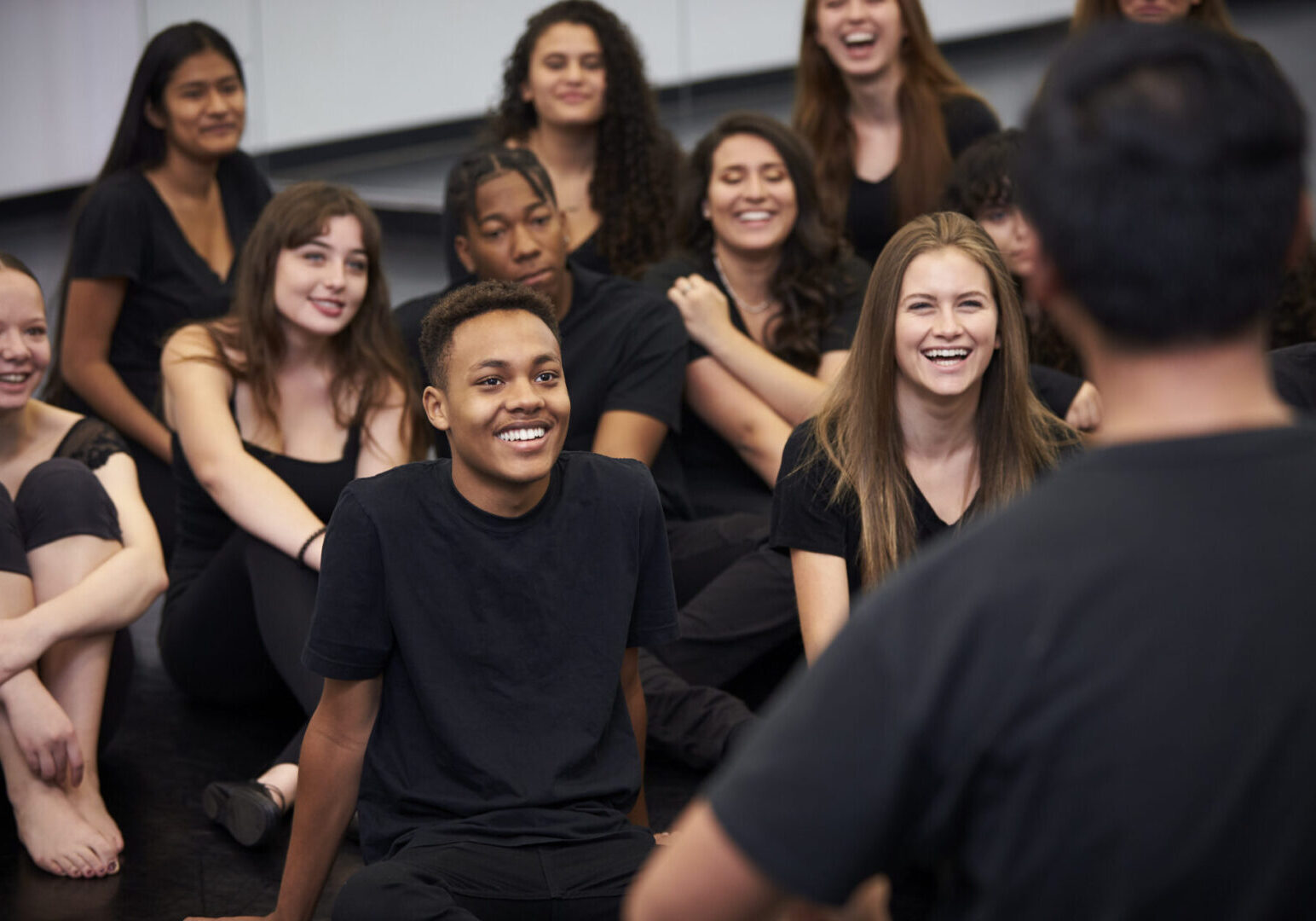 Teacher At Performing Arts School Talking To Students Sitting On Floor In Rehearsal Studio Group of people sitting and smiling together.